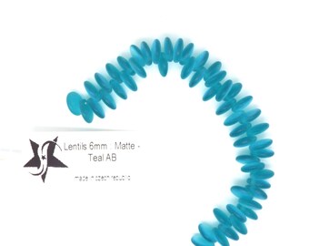 May include: A strand of teal blue glass lentil beads, 6mm in size, with a matte finish. The beads are arranged in a semi-circle shape. The text "Lentils 6mm Matte - Teal AB" is visible on a white label.