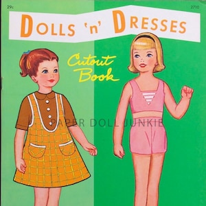 May include: A vintage paper doll book featuring two dolls, one with a yellow and brown plaid dress and the other with a pink top and shorts. The book cover has the title "Dolls 'n' Dresses Cutout Book" and the text "Paper Doll Junkie".