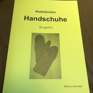 Könnte beinhalten: Ein gelbes Buch mit dem Titel "Nadelbinden Handschuhe So geht's" von Maria Lind Heel, mit einem Foto von einem Paar gestrickter Fäustlinge.