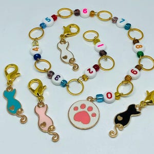 Könnte beinhalten: Ein Set aus bunten Katzen- und Pfoten-Charm-Maschenmarkierern mit goldenen Ringen und Perlen. Die Markierer sind von 0 bis 8 nummeriert.