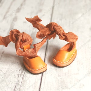 Pu&ograve; includere: Un paio di sandali in pelle arancione in miniatura con lacci in nastro arancione legati in un fiocco. I sandali sono su una superficie di legno bianco.