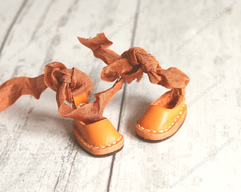 Pu&ograve; includere: Un paio di sandali in pelle arancione in miniatura con lacci in nastro arancione legati in un fiocco. I sandali sono su una superficie di legno bianco.