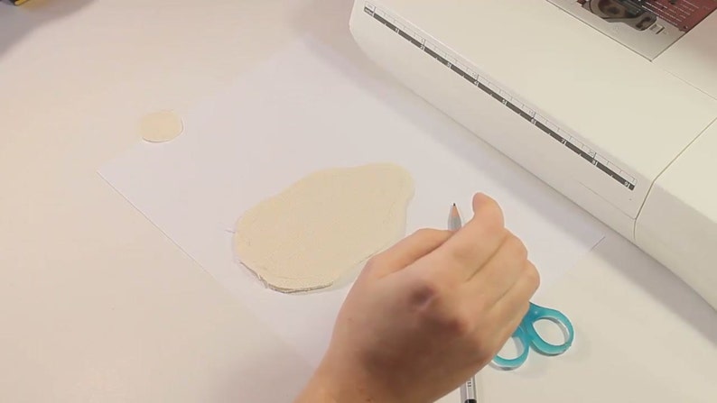 May include: A person is tracing the outline of a fabric shape with a pencil on a white surface. There is a smaller circle of fabric to the left of the larger shape.