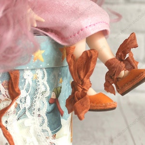 Pu&ograve; includere: Un paio di scarpe di sparto arancioni con lacci di nastro marrone. Le scarpe sono legate con un fiocco intorno alle caviglie. Le scarpe sono indossate da una bambola con i capelli rosa.