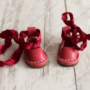 Pu&ograve; includere: Un paio di scarpe da bambola in pelle rossa con cuciture bianche e lacci in raso rosso.