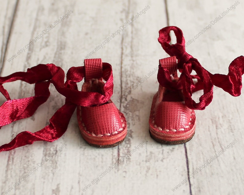 Pu&ograve; includere: Un paio di scarpe da bambola in pelle rossa con cuciture bianche e lacci in raso rosso.
