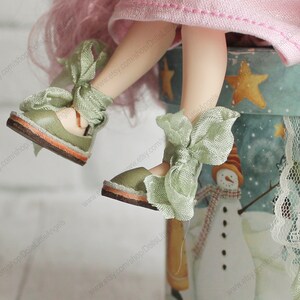 Pu&ograve; includere: Un paio di scarpe da bambola verdi con suole marroni chiare e fiocchi di raso verde. Le scarpe sono indossate da una bambola con i capelli rosa e un vestito rosa. La bambola &egrave; seduta su un contenitore blu e bianco con un disegno di pupazzo di neve.