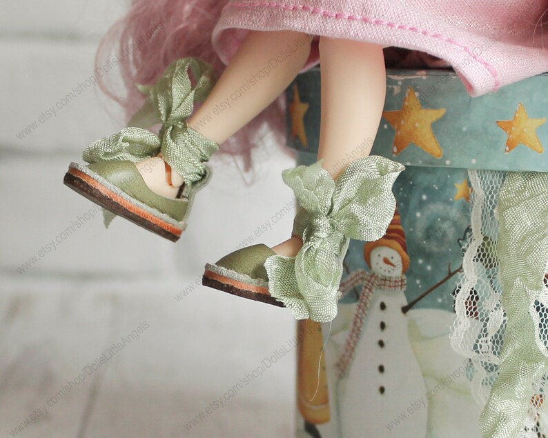 Pu&ograve; includere: Un paio di scarpe da bambola verdi con suole marroni chiare e fiocchi di raso verde. Le scarpe sono indossate da una bambola con i capelli rosa e un vestito rosa. La bambola &egrave; seduta su un contenitore blu e bianco con un disegno di pupazzo di neve.