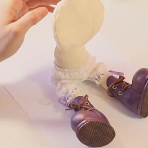 May include: A handmade doll with a white fabric body, white lace trim, and purple boots with brown laces. The doll is sitting on a white surface.