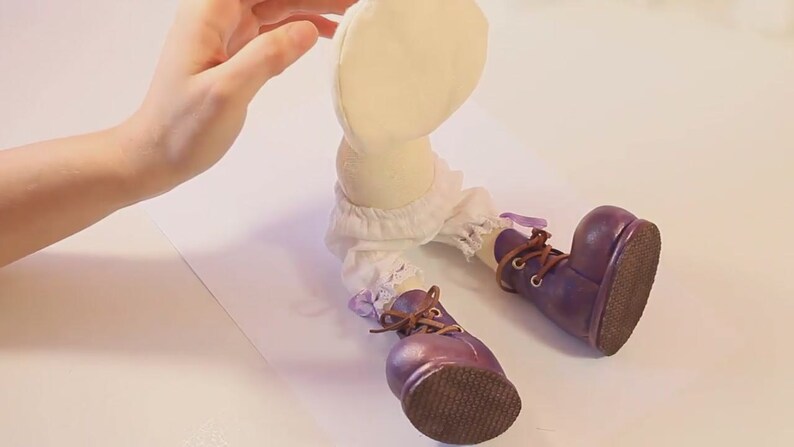 May include: A handmade doll with a white fabric body, white lace trim, and purple boots with brown laces. The doll is sitting on a white surface.