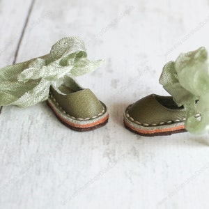 Pu&ograve; includere: Un paio di scarpe da bambola in pelle verde con un nastro verde legato intorno alle caviglie. Le scarpe hanno un bordo marrone e arancione intorno alla suola.