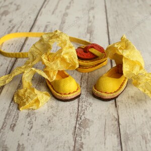 Pu&ograve; includere: Un paio di scarpe di bambola in pelle gialla con un nastro giallo legato intorno. Le scarpe hanno una borsa a forma di cuore rosso e giallo legata al nastro.