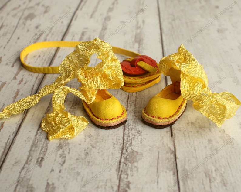 Pu&ograve; includere: Un paio di scarpe di bambola in pelle gialla con un nastro giallo legato intorno. Le scarpe hanno una borsa a forma di cuore rosso e giallo legata al nastro.