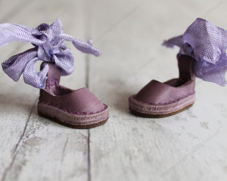 Pu&ograve; includere: Un paio di scarpe da bambola in pelle viola con un nastro viola legato intorno alla caviglia.