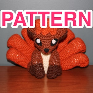 Könnte beinhalten: Gehäkelter Fuchs-Plüsch mit neun orangefarbenen Schwänzen, braunem Körper und weißen Akzenten. Das Wort "PATTERN" wird in rosa Buchstaben angezeigt. Der Fuchs hat große weiße Augen und eine schwarze Nase.