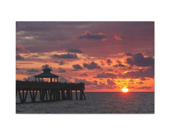 Könnte beinhalten: Leinwanddruck eines Sonnenuntergangs über dem Meer. Der Himmel ist mit orangefarbenen, roten und violetten Wolken gefüllt. Ein Holzsteg erstreckt sich in das Wasser, mit einem Pavillon am Ende. Die Sonne geht am Horizont unter.