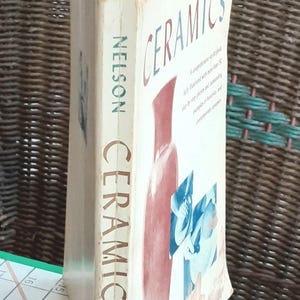 May include: A hardcover book titled "CERAMICS" by Nelson, with a red and blue illustration of pottery on the cover. The book's spine displays the title vertically. The book is open, revealing white pages.