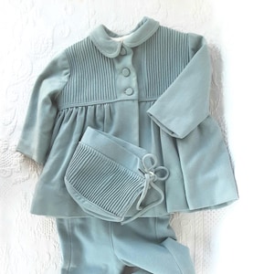 May include: A light blue baby outfit set, including a jacket, pants, and bonnet. The jacket has a pleated bodice and three buttons. The pants have flared bottoms. The bonnet has a pleated front and ties. The set is made of a soft fabric.