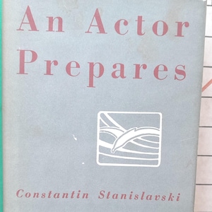Può includere: Un libro vintage intitolato "An Actor Prepares" di Constantin Stanislavski. La copertina è di un grigio-blu tenue con scritte rosse. Il libro presenta una grafica quadrata bianca e il testo "INTRODUCTION BY JOHN GIELGUD".