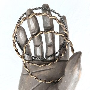 May include: Three metal bangles in varying designs. One bangle is made of small, dark beads. The other two are twisted metal, one in a gold tone and the other in a copper tone. The bangles are displayed on a grey hand sculpture against a white background.