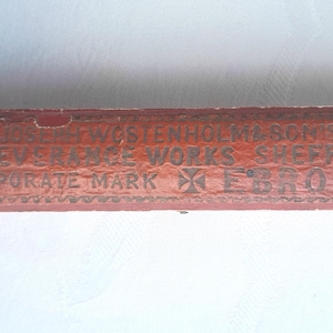 May include: A rectangular, antique wooden box with faded red paint. The box features gold lettering that reads "JOSEPH WESTENHOLM & SON'S PERSEVERANCE WORKS SHEFFIELD CORPORATE MARK EBRO". The box has a worn, vintage appearance.