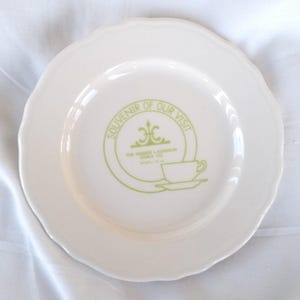 May include: Cream-coloured ceramic plate with a scalloped edge. The centre has a green circular design with the text "Souvenir of Our Visit" and a teacup illustration. The text "The Homer Laughlin China Co. Newell, W. Va." is also included.