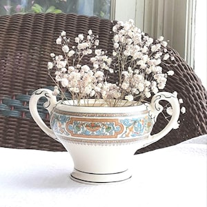Può includere: Un vaso in ceramica bianca con due manici, decorato con un motivo floreale colorato. Il vaso è pieno di fiori bianchi secchi. Il vaso è appoggiato su una superficie bianca.