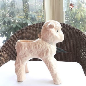 May include: A ceramic lamb planter in a soft, creamy pink. The lamb has a textured, wool-like appearance and a small opening on its back for planting. The planter is standing on four legs and has a detailed face.