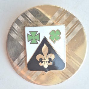 May include: A gold-toned round pin with a shield-shaped emblem. The emblem features a green cross, a green shamrock, and a black and gold fleur-de-lis on a white background. The pin has a textured, patterned border.