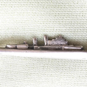 Puede incluir: Una figurita de barco de metal plateado. El barco en miniatura presenta estructuras detalladas y un casco largo y aerodinámico. El barco está sobre un fondo texturizado de color verde claro.