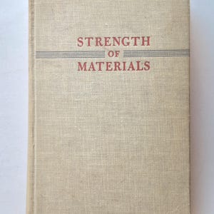 May include: A vintage book titled "Strength of Materials" with a beige linen cover. The title is in red letters with a gray horizontal line above and below the word "of". The book is closed and upright.