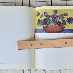 May include: An open book with a colorful illustration of a floral arrangement. The flowers, in shades of purple, blue, and yellow, are arranged in a red bowl. The background is yellow and blue.