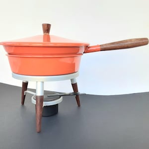 May include: An orange fondue pot with a wooden handle and lid knob. The pot sits on a silver stand with wooden legs. A small black burner is beneath the pot, ready for use. A vintage kitchenware item.