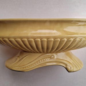 Puede incluir: Un macetero de cerámica amarillo con un diseño acanalado y una base decorativa. El macetero es perfecto para exhibir plantas o flores.