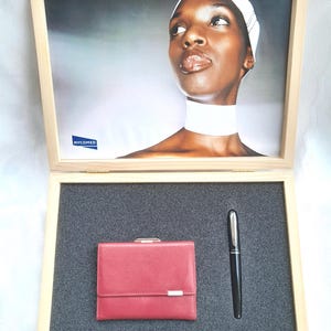 Puede incluir: Una caja de madera con una imagen enmarcada de una mujer con un pañuelo y un cuello blancos. Dentro, una cartera de cuero roja con un cierre plateado y un bolígrafo negro se exhiben sobre una superficie gris.