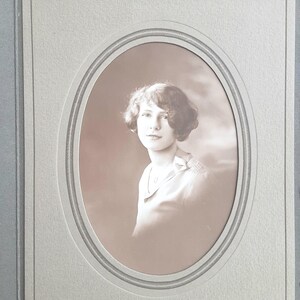 May include: Sepia-toned portrait of a woman in a light blouse, framed within an oval mount. The woman has short, wavy hair and is looking towards the viewer. The photograph is mounted in a light grey album.