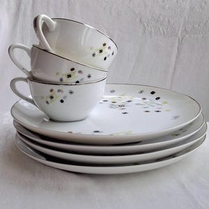 Peut inclure: Ensemble de tasses et d'assiettes en céramique blanche. Les tasses ont des anses et sont décorées de petits motifs abstraits colorés et de bordures argentées. Les assiettes sont empilées en dessous et ont des motifs assortis.