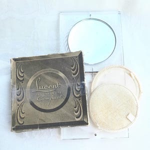 May include: Vintage Lucent Genuine Lucite Compact with a mirror and two powder puffs. The compact box is square with decorative scrollwork and the text "Lucent Genuine Lucite Compact". The mirror is round and set in a clear frame.