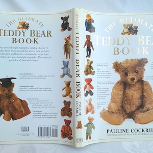 May include: Open book titled "The Ultimate Teddy Bear Book" with a brown teddy bear on the right page. The left page features various teddy bear illustrations. The book cover has gold lettering and illustrations of teddy bears.