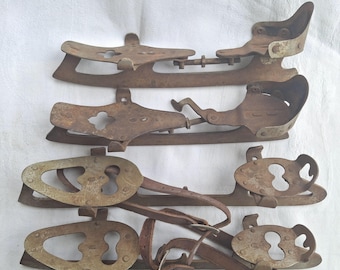 May include: Four pairs of antique ice skates, showing signs of age and wear. The skates are made of metal and leather, with a rusty brown patina. The blades are attached to the shoe supports with metal hardware.
