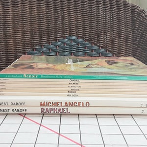 May include: A stack of art books featuring titles like "Renoir," "Chagall," and "Michelangelo." The books have white spines with titles in various colors. The background includes a wicker chair and a checkered surface.