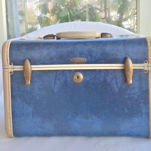 Könnte beinhalten: Ein blauer Vintage-Koffer mit cremefarbenen Zierleisten und einem passenden Griff. Der Koffer hat messingfarbene Beschläge, einschließlich Verschlüssen und einem Schlüsselloch. Das Äußere hat ein strukturiertes Aussehen, und das Design deutet auf eine klassische Reiseästhetik hin.