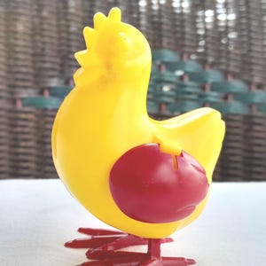 Könnte beinhalten: Ein hellgelbes Plastikspielzeug in Form eines Huhns mit einem roten, kreisförmigen Teil an der Seite. Das Huhn hat rote Füße und einen Kamm. Das Spielzeug steht auf einer weißen Oberfläche.