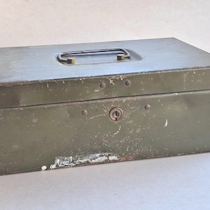 May include: A rectangular, olive-green metal box with a handle on the lid. The box has a lock and keyhole on the front. The paint is worn, showing signs of age and use. The box is likely used for storage.