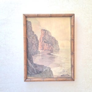 May include: A framed print depicting a coastal scene with prominent rock formations and a sandy beach. The artwork is encased in a bamboo-style frame. The color scheme includes beige, grey, and pink tones, evoking a tranquil, natural atmosphere.