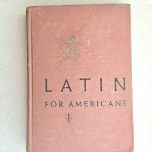 May include: A vintage book titled "LATIN FOR AMERICANS" in a muted rose colour. The cover features a gold illustration of a running figure and a torch. The book's title is printed in a bold, black font.