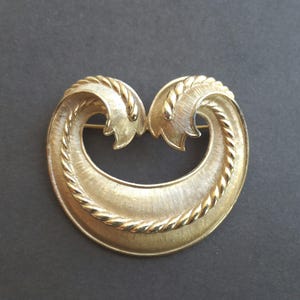 May include: A gold-toned vintage brooch with a heart-shaped design. The brooch features a textured surface with a rope-like border and leaf-shaped accents. The pin is set against a dark background.