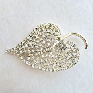 May include: A silver-toned leaf-shaped brooch, encrusted with numerous clear rhinestones. The brooch features a delicate, openwork design, with a curved stem detail. The overall aesthetic is elegant and suitable for formal wear.