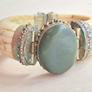 May include: A hinged bracelet with a large oval green stone centerpiece. The bracelet has a cream-coloured, segmented band and is accented with silver-toned metal and small decorative beads. The bracelet is approximately 5 cm wide.