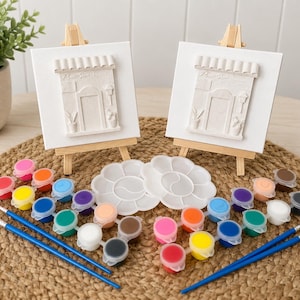 Könnte beinhalten: Zwei weiße quadratische Leinwände mit einem Relief einer Ladenfassade, die jeweils auf einer kleinen Holzstaffelei stehen. Das Set enthält zwei weiße blumenförmige Farbpaletten, mehrere kleine Töpfe mit Acrylfarben und zwei Pinsel.